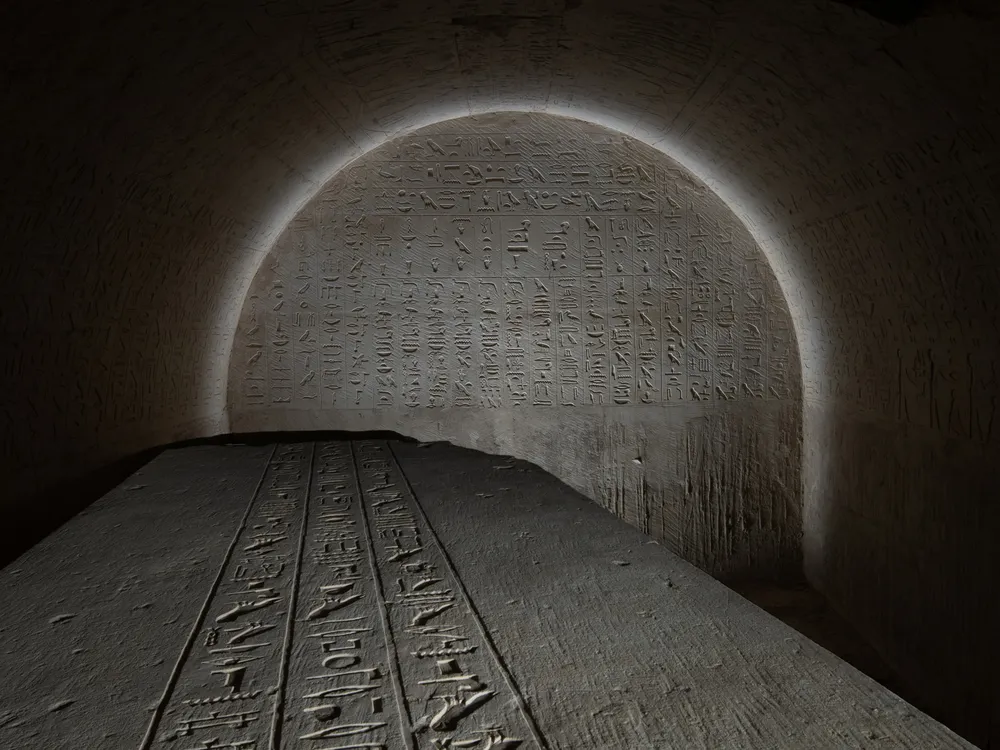 the top of a sarcophagus covered in writing with a glyph-covered wall behind it lit in an arch