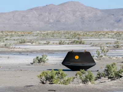 A black, saucer-like capsule sits on the desert surface