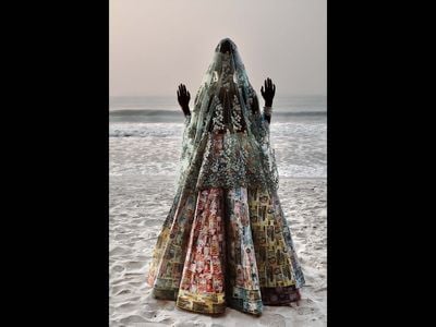 a person stands  in a decorated dress and vail on a beach with the sea behind them