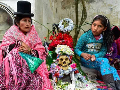 A woman in traditional Aymara dress sits with her daughter and their honored human skull, or ñatita, and a bag of coca leaves during the 2015 Fiesta de las Ñatitas in Bolivia.